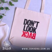 Шопер не дивись назад. Бежевий колір, ракурс зверху, фон світлий. Тканина середньої товщини, ручки середньої довжини. Місткість достатня для повсякденних речей, принт з написом 'DON'T LOOK BACK' в чорному та червоному кольорах.
