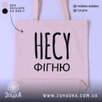 Шопер несу фігню, бежевий, зображений з фронтального ракурсу на світлому фоні, має великий чорний напис 'НЕСУ ФІГНЮ', з двома ручками, прямокутна форма, місткість достатня для повсякденних речей.