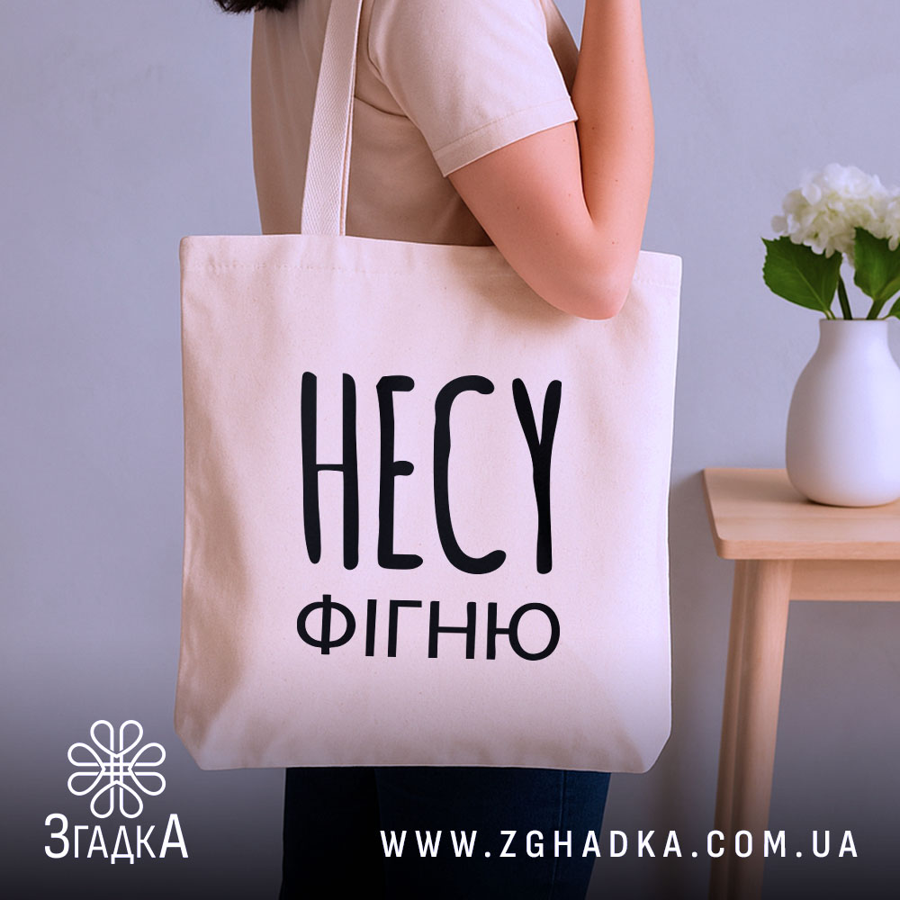 Шопер несу фігню, бежевий, зображений з боку, на фоні нейтральної стіни, великий чорний напис 'НЕСУ ФІГНЮ', з двома ручками, прямокутна форма, місткість підходить для покупок.