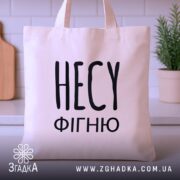 Шопер несу фігню, бежевий, зображений з фронтального ракурсу на фоні кухонних елементів, великий чорний напис 'НЕСУ ФІГНЮ', з двома ручками, прямокутна форма, місткість достатня для продуктів.