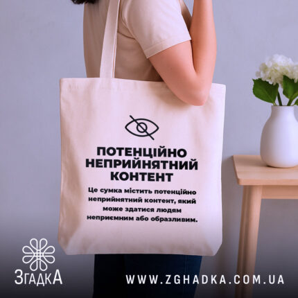 Шопер потенційно неприйнятний контент. Сумка бежевого кольору, зображена на плечі людини, фон з елементами інтер'єру. Тканина середньої товщини, ручки довгі, місткість достатня для покупок, принт чорного кольору з текстом на передній частині.