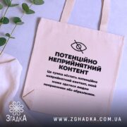 Шопер потенційно неприйнятний контент. Сумка бежевого кольору, зображена на столі поруч з рослинами. Тканина середньої товщини, ручки середньої довжини, місткість достатня для повсякденних речей, принт чорного кольору з текстом на передній частині.