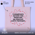 Шопер святковий настрій, бежевий, з фронтальної сторони, на нейтральному фоні, з тонкої бавовняної тканини, ручки середньої довжини, місткий, з принтом у вигляді червоних гірлянд навколо тексту.
