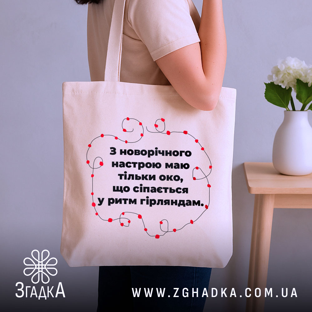 Шопер святковий настрій, бежевий, з бокового ракурсу, на фоні кімнати з рослиною, з тонкої бавовняної тканини, ручки середньої довжини, місткий, з принтом у вигляді червоних гірлянд навколо тексту.