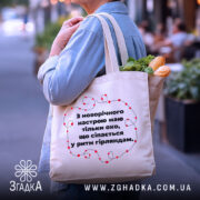 Шопер святковий настрій, бежевий, з бокового ракурсу, на фоні вулиці, з тонкої бавовняної тканини, ручки середньої довжини, місткий, з принтом у вигляді червоних гірлянд навколо тексту.