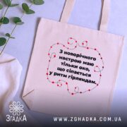 Шопер святковий настрій, бежевий, з фронтальної сторони, на фоні текстильних виробів, з тонкої бавовняної тканини, ручки середньої довжини, місткий, з принтом у вигляді червоних гірлянд навколо тексту.