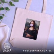 Шопер жартівлива Мона Ліза, бежевий, з верхнього ракурсу, на фоні столу з рослинами, з тонкої бавовняної тканини, з короткими ручками, місткий, з принтом класичної картини Мона Ліза з жувальною гумкою в роті.