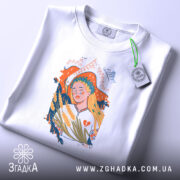 Футболка Українська дівчина білого кольору, складена на поверхні, ракурс зверху, фон світло-сірий, принт з дівчиною з вінком на голові, що тримає колосся, з етикеткою зеленого кольору.