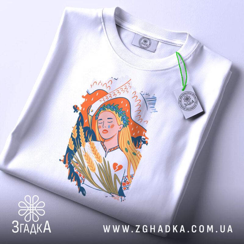 Футболка Українська дівчина білого кольору, складена на поверхні, ракурс зверху, фон світло-сірий, принт з дівчиною з вінком на голові, що тримає колосся, з етикеткою зеленого кольору.