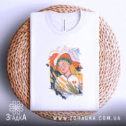 Футболка Українська дівчина білого кольору, складена на кошику, ракурс зверху, фон світло-сірий, принт з дівчиною з вінком, що тримає колосся, на плетеній основі.