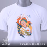 Футболка Українська дівчина білого кольору, складена на поверхні, ракурс зверху, фон світло-сірий, принт з дівчиною з вінком на голові, що тримає колосся, з етикеткою зеленого кольору.