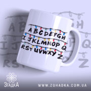 Чашка алфавіт з гірляндою, біла, зображена з правого ракурсу, фон світло-фіолетовий, принт з буквами A-Z, обрамленими кольоровими гірляндами, ручка кругла, край рівний, глянцева поверхня.