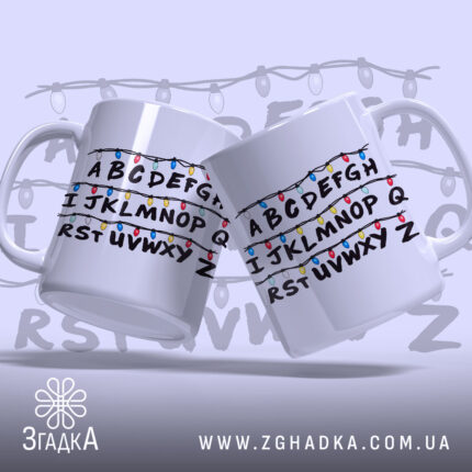 Чашка алфавіт з гірляндою, біла, зображена з двох боків, фон світло-фіолетовий, принт з буквами A-Z, обрамленими кольоровими гірляндами, ручки круглі, краї рівні, глянцева поверхня.
