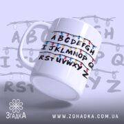 Чашка алфавіт з гірляндою, біла, зображена під кутом, фон світло-фіолетовий, принт з буквами A-Z, обрамленими кольоровими гірляндами, ручка кругла, край рівний, глянцева поверхня.