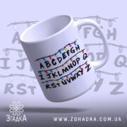 Чашка алфавіт з гірляндою, біла, зображена з правого ракурсу, фон світло-фіолетовий, принт з буквами A-Z, обрамленими кольоровими гірляндами, ручка кругла, край рівний, глянцева поверхня.