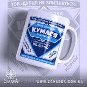 Чашка для Кумасі, біла з синім принтом, зображена під кутом, на фоні з розмитим логотипом. Принт містить текст та графічні елементи, ручка класична, край рівний, поверхня глянцева.