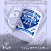 Чашка для Кумасі, біла з синім принтом, зображена збоку, на фоні з розмитим логотипом. Принт містить текст та графічні елементи, ручка класична, край рівний, поверхня глянцева.