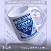 Чашка для Кумасі, біла з синім принтом, зображена під кутом, на фоні з розмитим логотипом. Принт містить текст та графічні елементи, ручка класична, край рівний, поверхня глянцева.