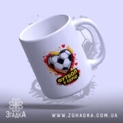 Чашка Футбол у серці, біла, з зображенням футбольного м'яча у формі серця, ручка стандартна, край рівний, фон з розмитими плямами червоного і жовтого кольору, глянцева поверхня.