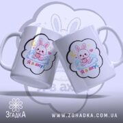 Чашка подарунок з зайчиком, дві чашки під кутом, біла з кольоровим принтом, зайчик на хмарі з чашкою, напис 'Я в ахуї' на кожній чашці, ручки класичні, край рівний, глянцева поверхня.