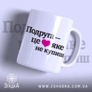 Чашка про дружбу подрузі, біла, з ракурсом спереду, на фоні світло-фіолетового кольору, з чорним написом "Подруга – це ❤️ яке не купиш", ручка круглої форми, край рівний, глянцева поверхня.