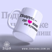 Чашка про дружбу подрузі, біла, з ракурсом знизу, на фоні світло-фіолетового кольору, з чорним написом "Подруга – це ❤️ яке не купиш", ручка круглої форми, край рівний, глянцева поверхня.