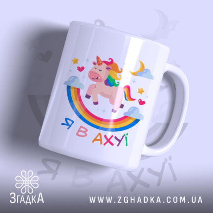 Чашка зі здивованим єдинорогом, білого кольору, з ракурсу спереду, на фоні світло-фіолетового кольору. Принт містить єдинорога, веселку, зірки та напис 'Я В АХУЙ'. Ручка чашки гладка, край рівний, поверхня глянцева.