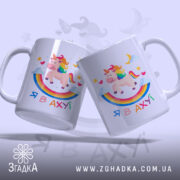 Чашка зі здивованим єдинорогом, білого кольору, з ракурсу, де видно дві чашки, на фоні світло-фіолетового кольору. Принт містить єдинорога, веселку, зірки та напис 'Я В АХУЙ'. Ручки чашок гладкі, краї рівні, поверхня глянцева.