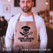 Фартух годую з любов’ю, бежевого кольору, на чоловікові, що позує на фоні кафе, принт з черепом та кухонними приладами розташований по центру.