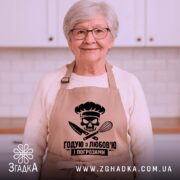Фартух годую з любов’ю, бежевого кольору, зображений на манекені з нейтральним фоном, принт з черепом та кухонними приладами розташований по центру.