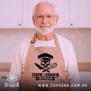 Фартух годую з любов’ю, бежевого кольору, зображений на дерев'яному фоні з елементами декору, принт з черепом та кухонними приладами розташований по центру.