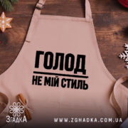 Фартух голод не мій стиль. Бежевий фартух, знятий зверху на дерев'яному фоні. На фоні присутні декорації у вигляді новорічних елементів. Принт чорного кольору з написом 'ГОЛОД НЕ МІЙ СТИЛЬ'.
