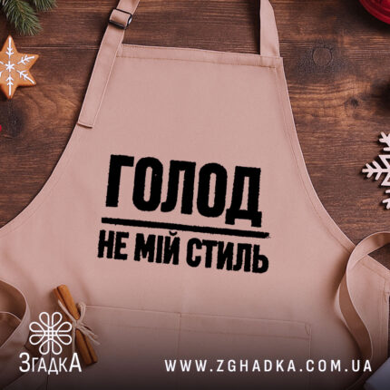 Фартух голод не мій стиль. Бежевий фартух, знятий зверху на дерев'яному фоні. На фоні присутні декорації у вигляді новорічних елементів. Принт чорного кольору з написом 'ГОЛОД НЕ МІЙ СТИЛЬ'.