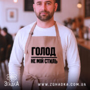 Фартух голод не мій стиль. Бежевий фартух, зображений на чоловікові, який стоїть у кафе. Фон розмитий, з елементами інтер'єру. Принт чорного кольору з написом 'ГОЛОД НЕ МІЙ СТИЛЬ'.