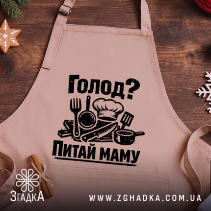 Фартух голод питай маму. Бежевий фартух, зображений з близького ракурсу на дерев'яному фоні. На фартусі чорний принт з кухонними приладами, прикрашений новорічними елементами.