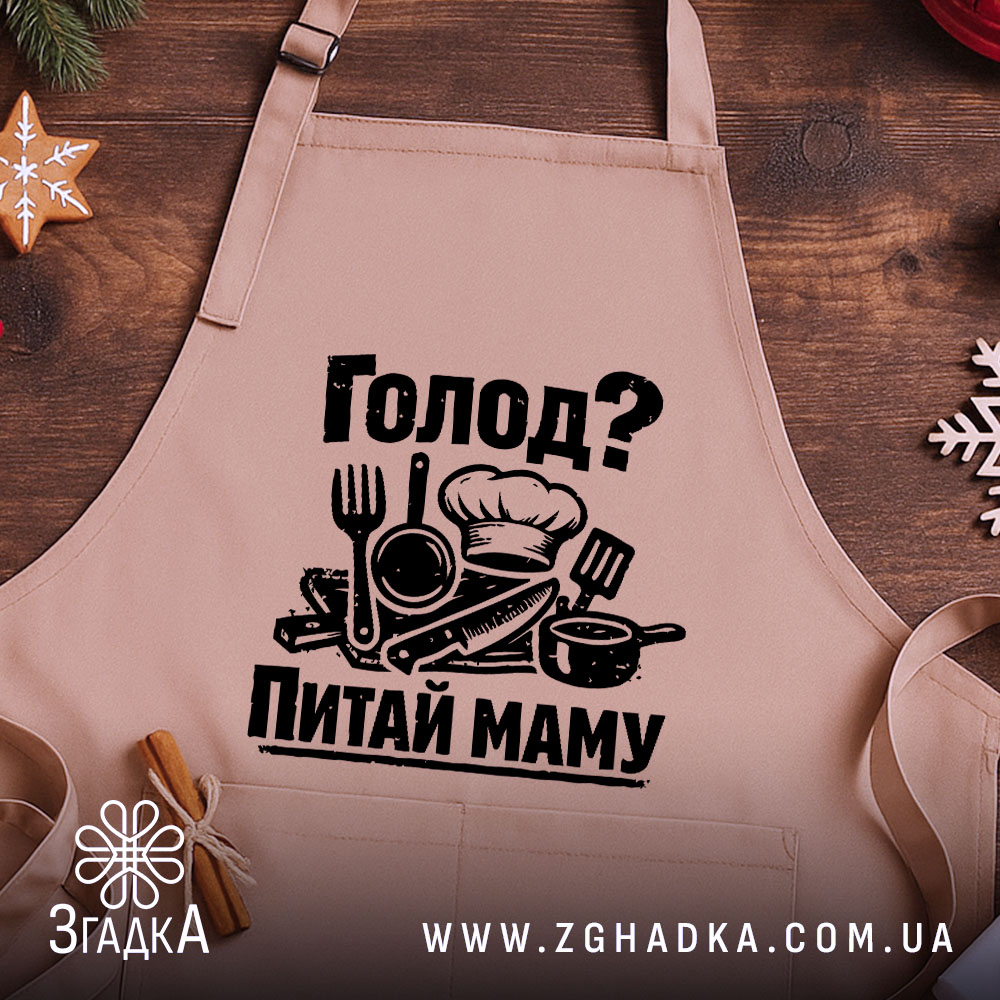 Фартух голод питай маму. Бежевий фартух, зображений з близького ракурсу на дерев'яному фоні. На фартусі чорний принт з кухонними приладами, прикрашений новорічними елементами.