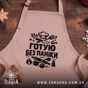 Фартух готую без паніки, бежевого кольору, зображений на дерев'яному фоні, принт чорного кольору з кухонними предметами, навколо святкові елементи.