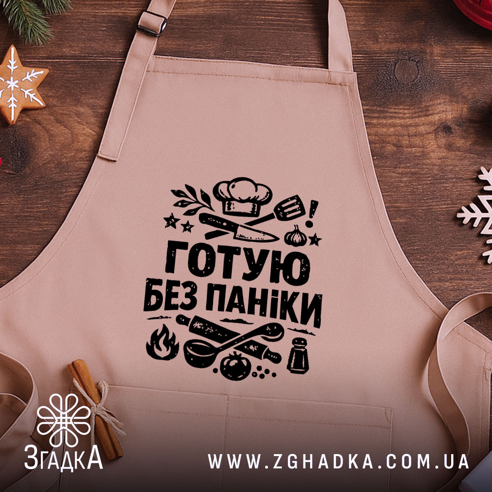 Фартух готую без паніки, бежевого кольору, зображений на дерев'яному фоні, принт чорного кольору з кухонними предметами, навколо святкові елементи.