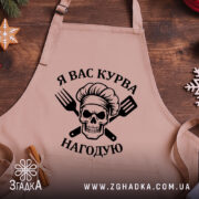 Фартух я вас курва нагодую, бежевого кольору, з близького ракурсу на дерев'яному фоні, принт з черепом та кухонними приладами чорного кольору, декор у вигляді новорічних елементів.