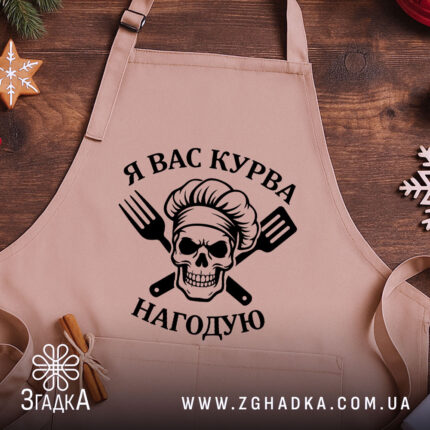 Фартух я вас курва нагодую, бежевого кольору, з близького ракурсу на дерев'яному фоні, принт з черепом та кухонними приладами чорного кольору, декор у вигляді новорічних елементів.