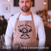 Фартух я вас курва нагодую, бежевого кольору, на чоловікові, фон кафе з розмитими фоновими елементами, принт з черепом та кухонними приладами чорного кольору.