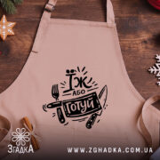 Фартух їж або готуй, бежевого кольору, зображений з близької відстані на дерев'яному фоні, принт чорного кольору з написом 'Їж або готуй' та зображенням виделки і ножа, поруч з декором у вигляді новорічних зірочок.