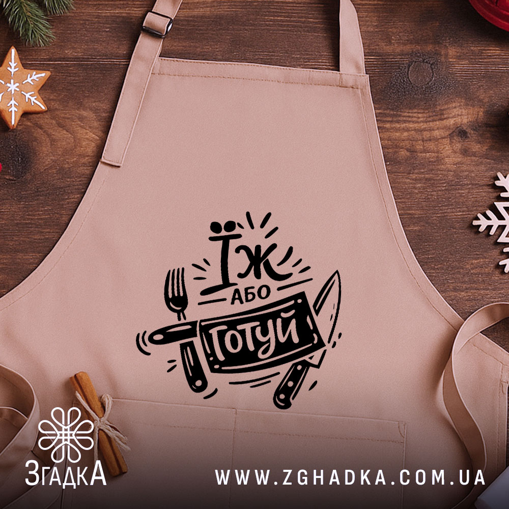 Фартух їж або готуй, бежевого кольору, зображений з близької відстані на дерев'яному фоні, принт чорного кольору з написом 'Їж або готуй' та зображенням виделки і ножа, поруч з декором у вигляді новорічних зірочок.