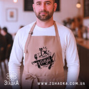 Фартух їж або готуй, бежевого кольору, зображений на чоловікові з бородою, фон кафе з розмитими фігурами людей, принт чорного кольору з написом 'Їж або готуй' та зображенням виделки і ножа.