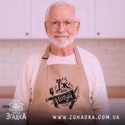 Фартух їж або готуй, бежевого кольору, зображений з близької відстані на дерев'яному фоні, принт чорного кольору з написом 'Їж або готуй' та зображенням виделки і ножа, поруч з декором у вигляді новорічних зірочок.