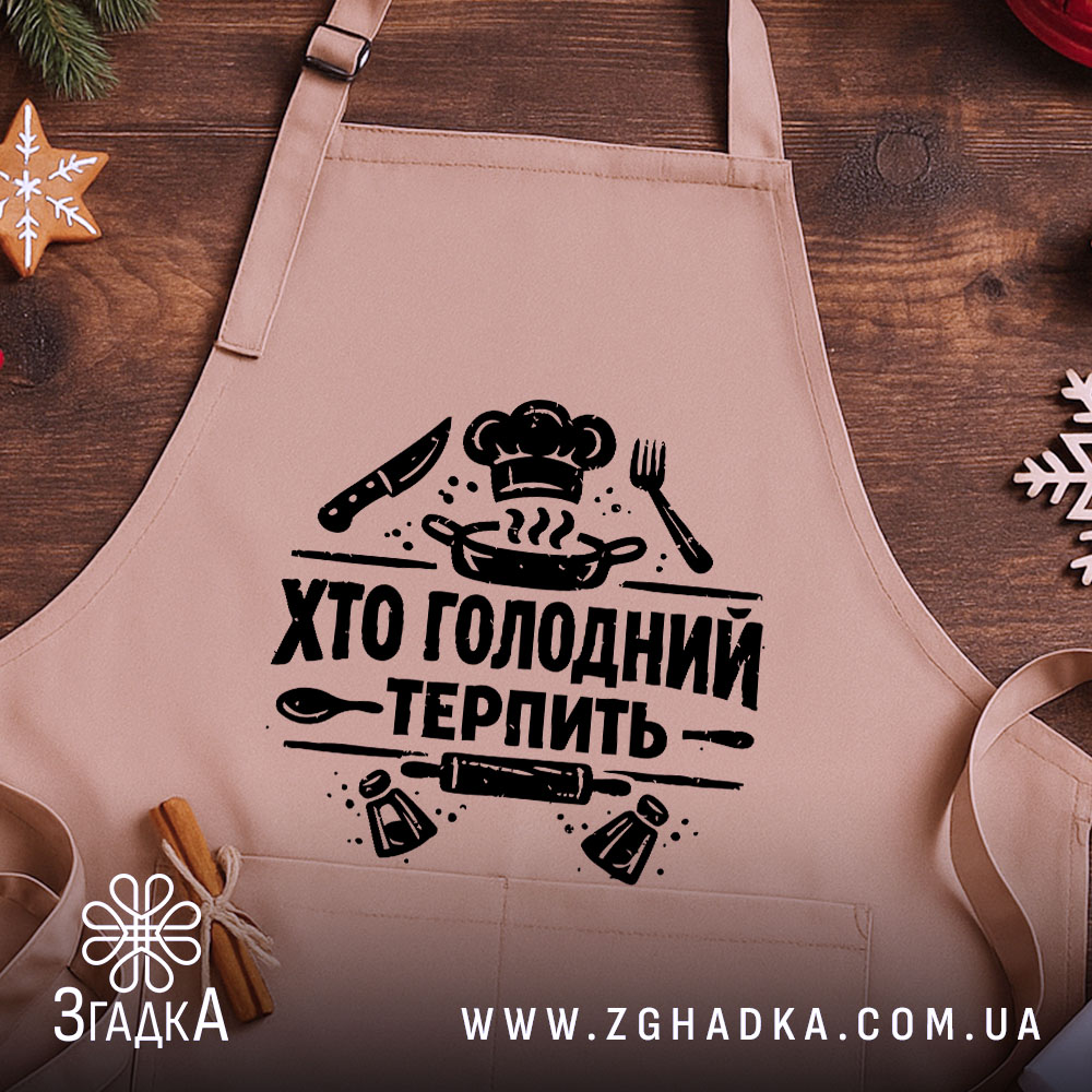 Фартух хто голодний, бежевого кольору, зображений на дерев'яному фоні, принт чорного кольору з написом 'Хто голодний терпіть' та кухонними атрибутами, на фоні видно святкові елементи.