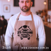 Фартух хто голодний, бежевого кольору, зображений на чоловікові в кафе, фон розмитий, принт чорного кольору з написом 'Хто голодний терпіть' та кухонними атрибутами.