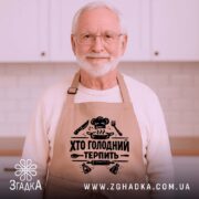 Фартух хто голодний, бежевого кольору, зображений на дерев'яному фоні, принт чорного кольору з написом 'Хто голодний терпіть' та кухонними атрибутами, на фоні видно святкові елементи.