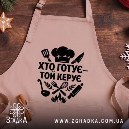 Фартух хто готує керує, бежевий колір, зображення з верхнього ракурсу, фон дерев'яний, принт чорного кольору з кухонними атрибутами.