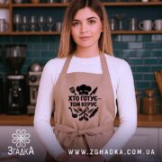Фартух хто готує керує, бежевий колір, зображення з фронтального ракурсу на жінці, фон кухонний з темно-зеленими елементами, принт чорного кольору з кухонними атрибутами.